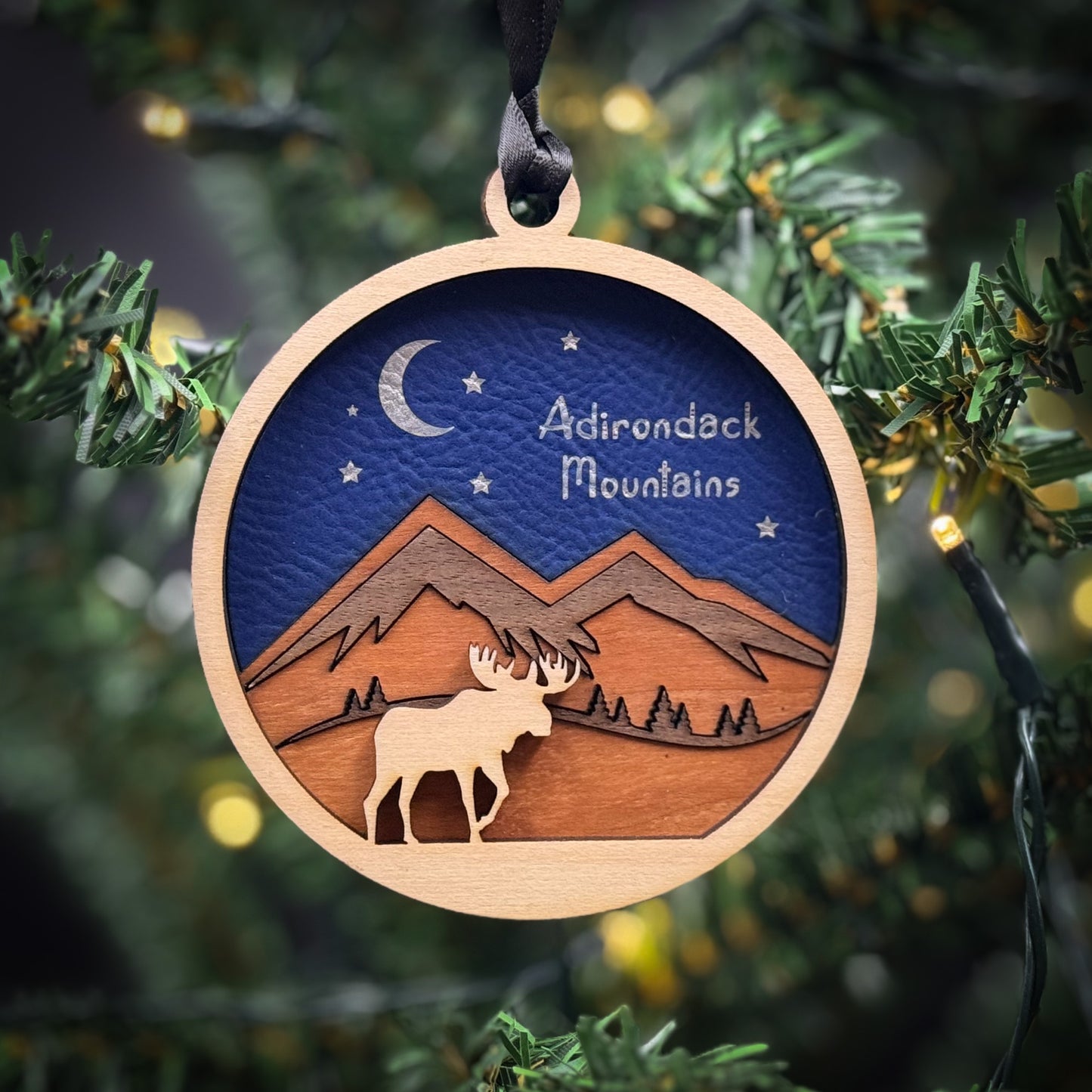 A wooden ornament hanging on a Christmas tree, depicting a moose silhouette against a night sky with mountains and stars.