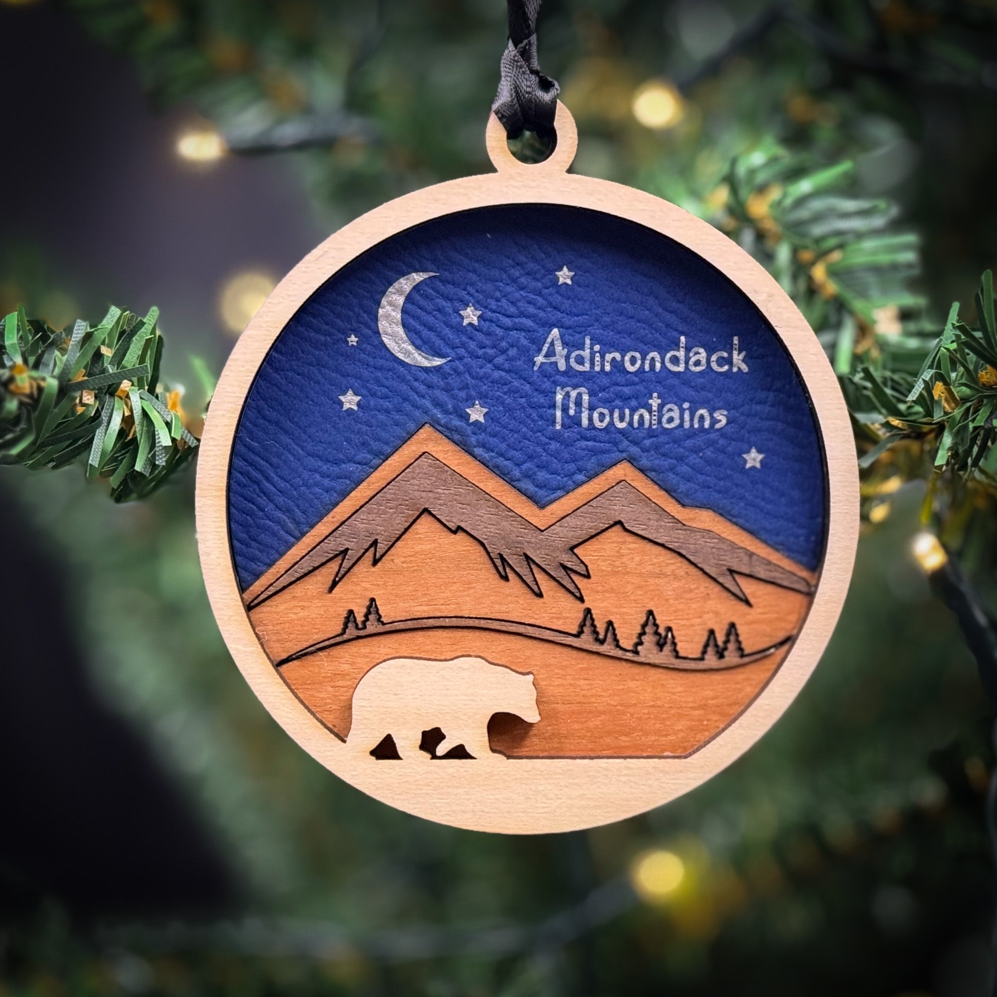 A wooden ornament hanging on a tree, depicting a bear silhouette against a night sky with mountains and stars.
