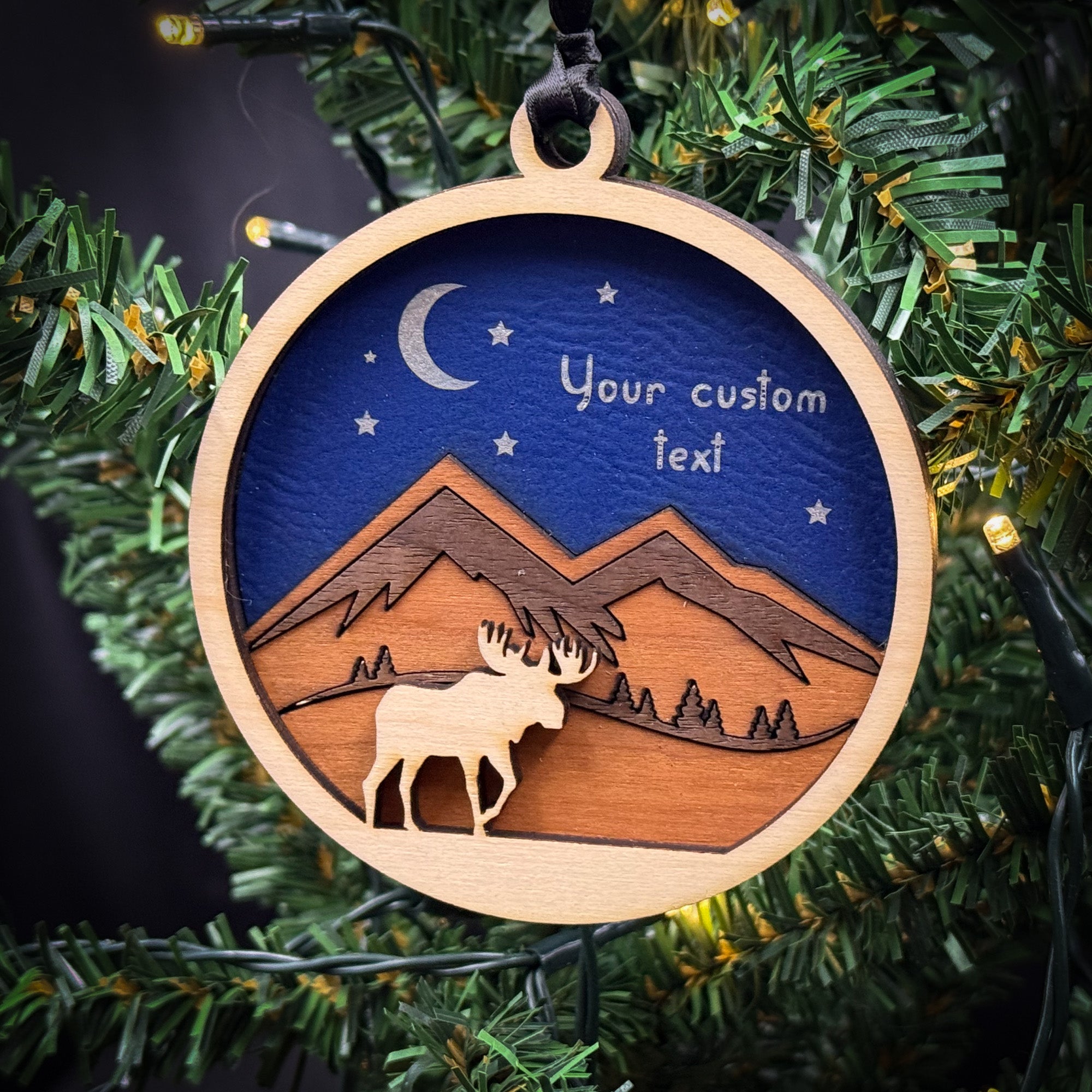 A wooden ornament hanging on a Christmas tree, depicting a moose silhouette against a night sky with mountains and stars.