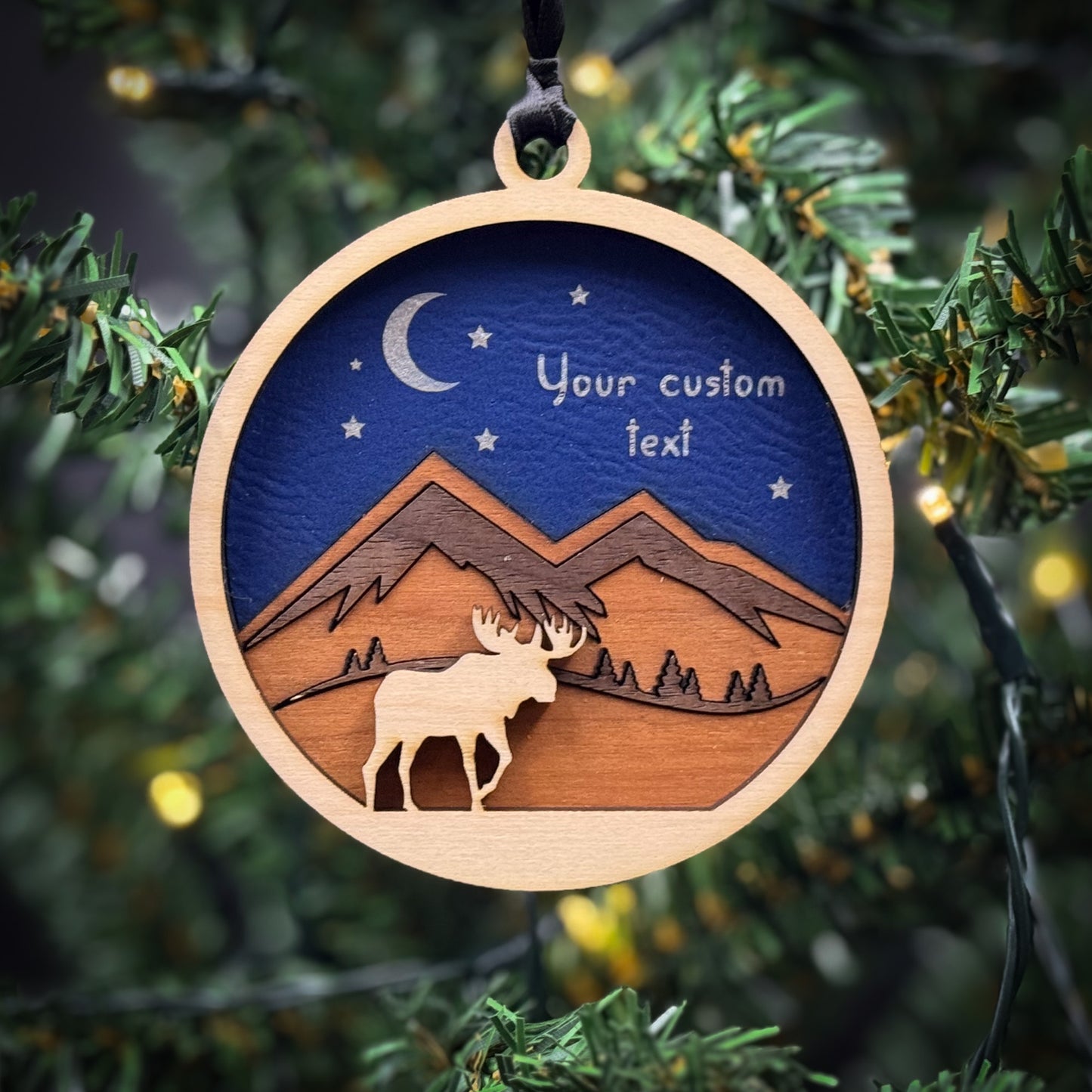 A wooden ornament with a moose silhouette and a mountain landscape, hanging on a Christmas tree.