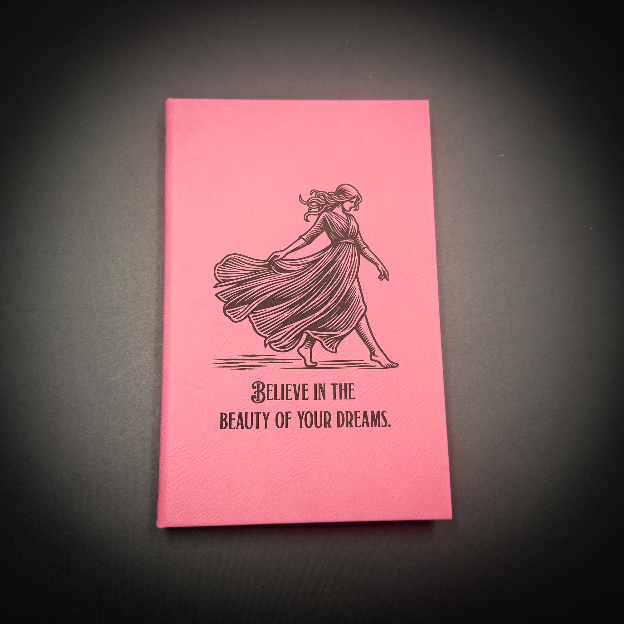 A pink notebook with a black illustration of a woman in a flowing dress and the text "Believe in the beauty of your dreams" printed on it.
