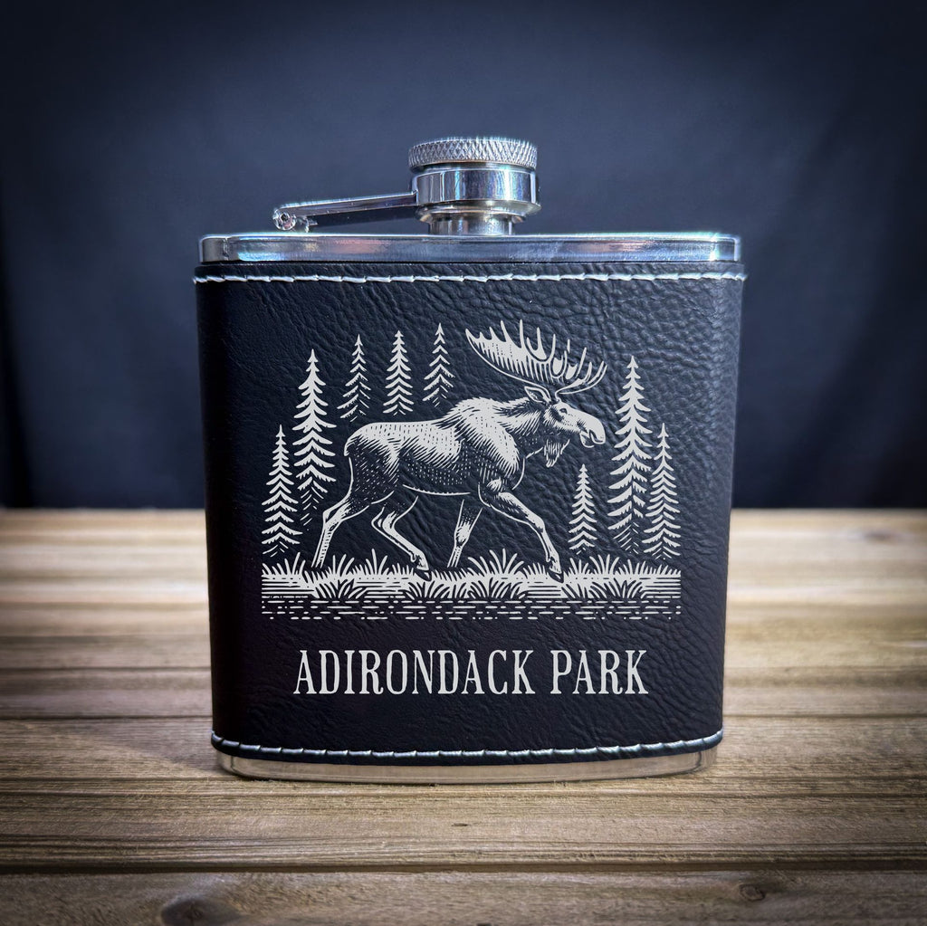 A black leather flask with a white illustration of a moose and pine trees on it, placed on a wooden surface.