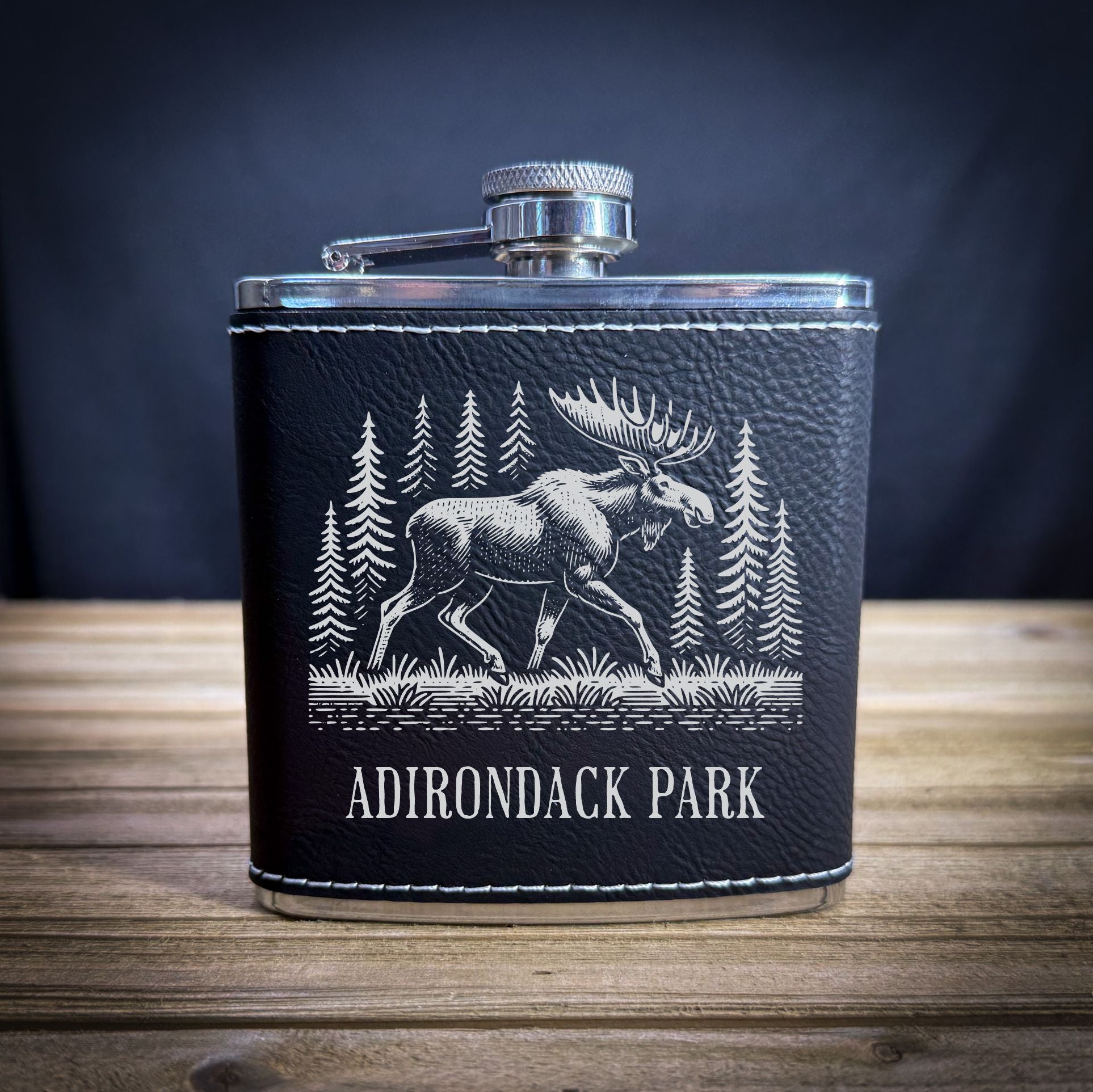 A black leather flask with a white illustration of a moose and pine trees on it, placed on a wooden surface.