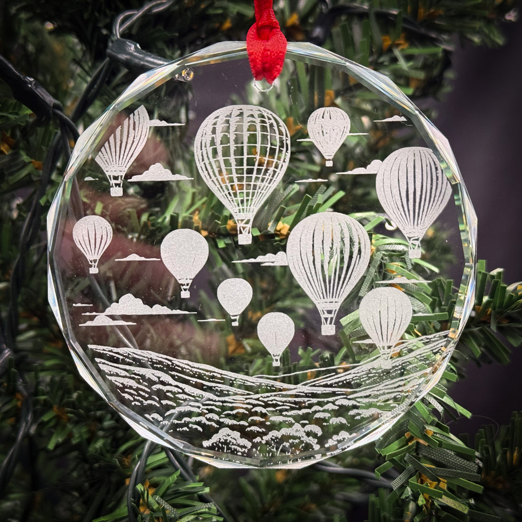 A clear glass ornament hanging on a Christmas tree, featuring a design of hot air balloons floating in the sky.