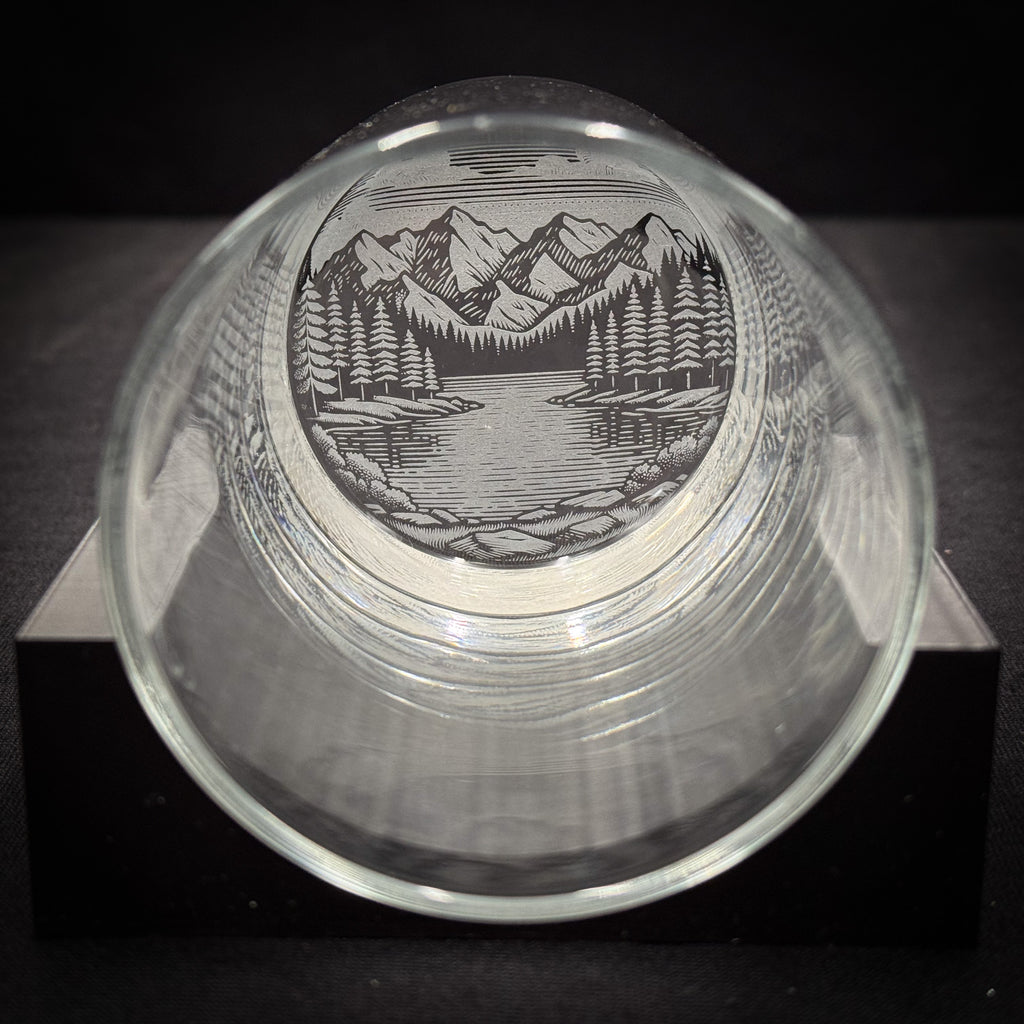 a glass with a mountain landscape etched onto it, sitting on a black surface.