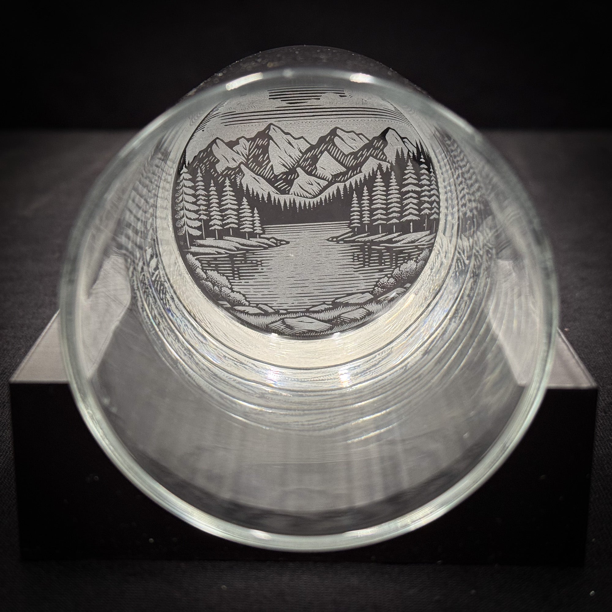 a glass with a mountain landscape etched onto it, sitting on a black surface.