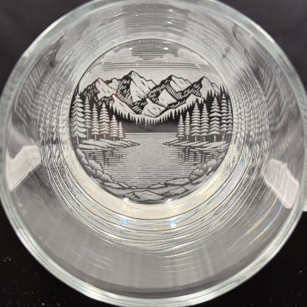 a glass with a black and white illustration of a mountain landscape, including a lake and pine trees, encased within the glass.