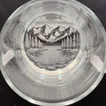 a glass with a black and white illustration of a mountain landscape, including a lake and pine trees, encased within the glass.