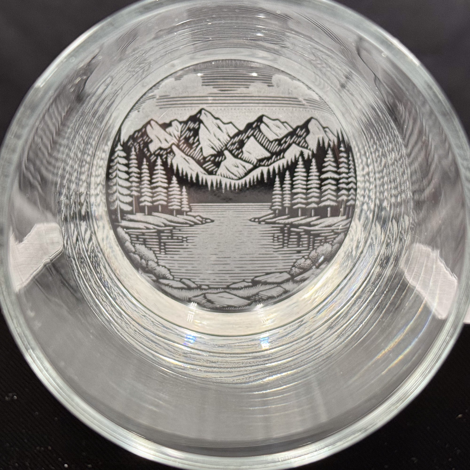 a glass with a black and white illustration of a mountain landscape, including a lake and pine trees, encased within the glass.