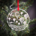 A clear glass ornament hanging from a tree branch, featuring a design of hot air balloons floating in the sky.