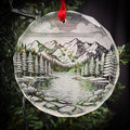 A clear glass ornament hanging on a tree, featuring a landscape scene with mountains, trees, and a lake.