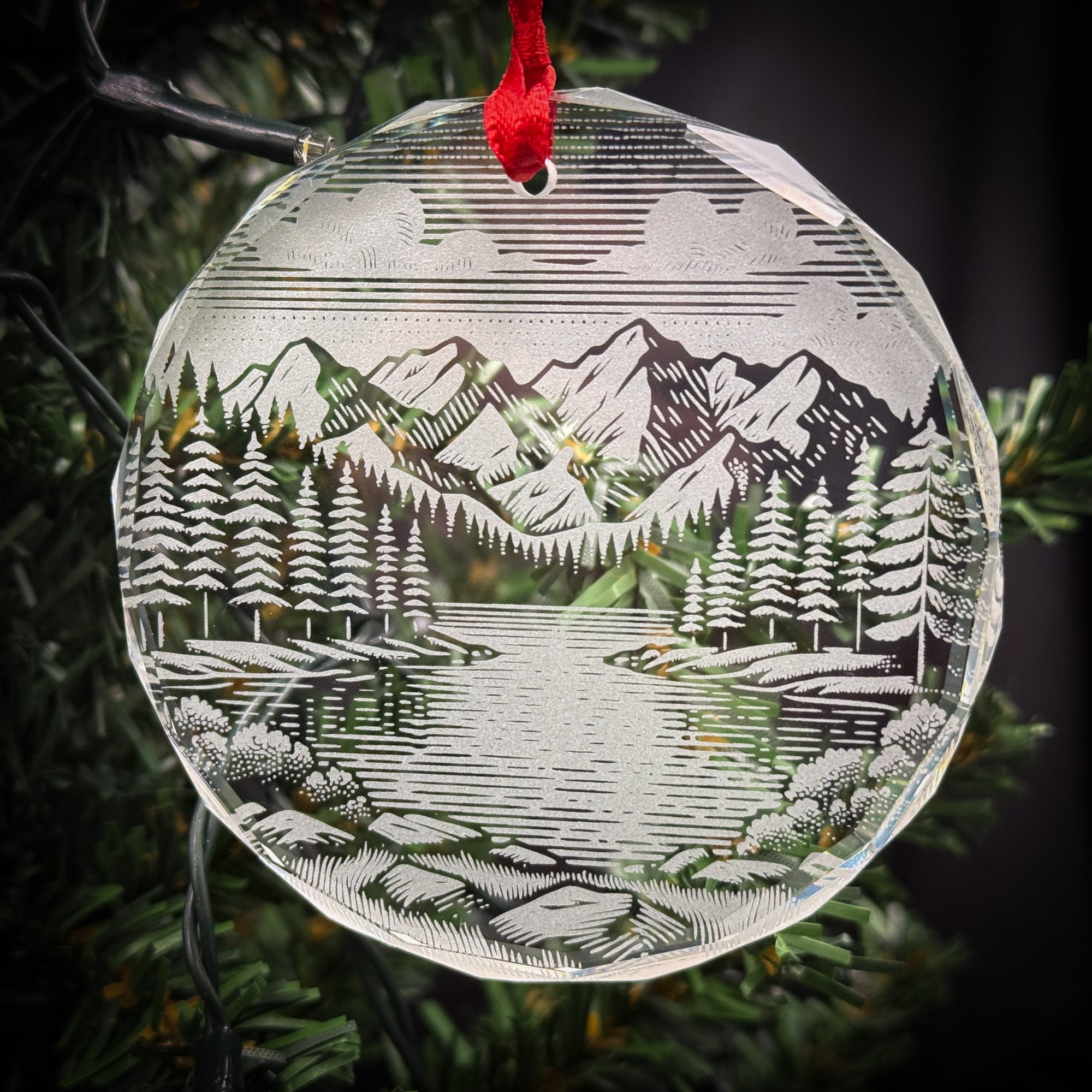 A clear glass ornament hanging on a tree, featuring a landscape scene with mountains, trees, and a lake.
