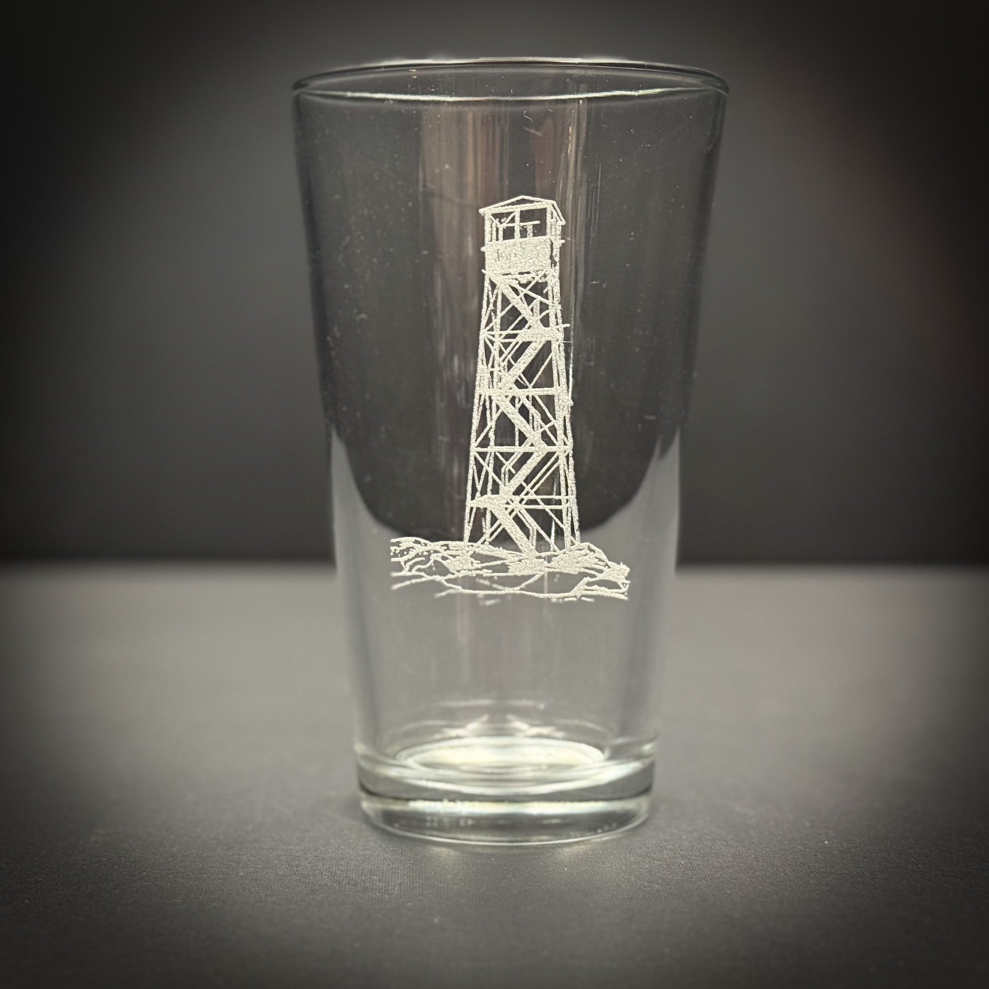 A clear glass with a white lighthouse design etched on it, sitting on a gray surface.