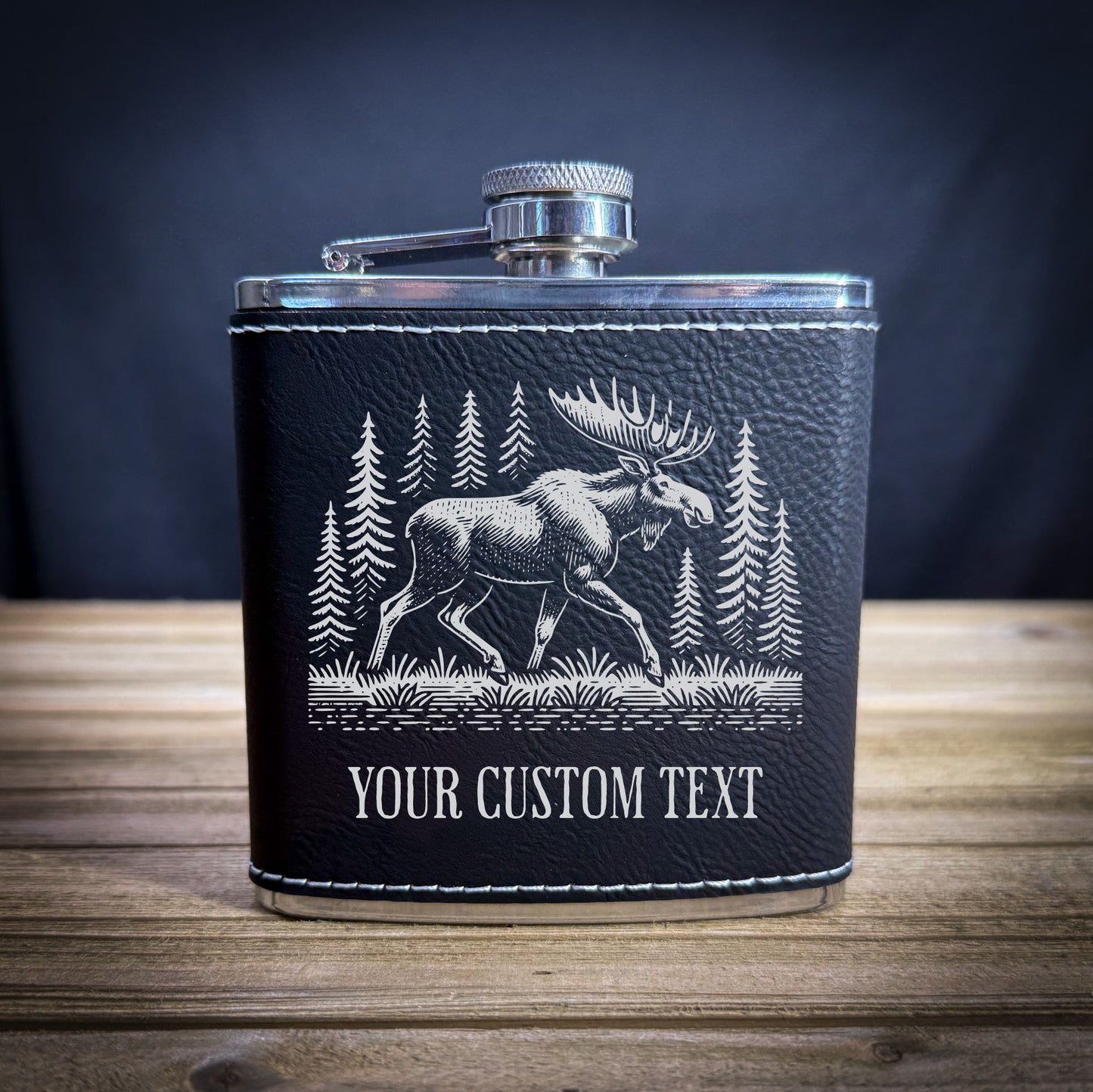 A black leather flask with a silver cap and a white illustration of a moose in a forest setting.