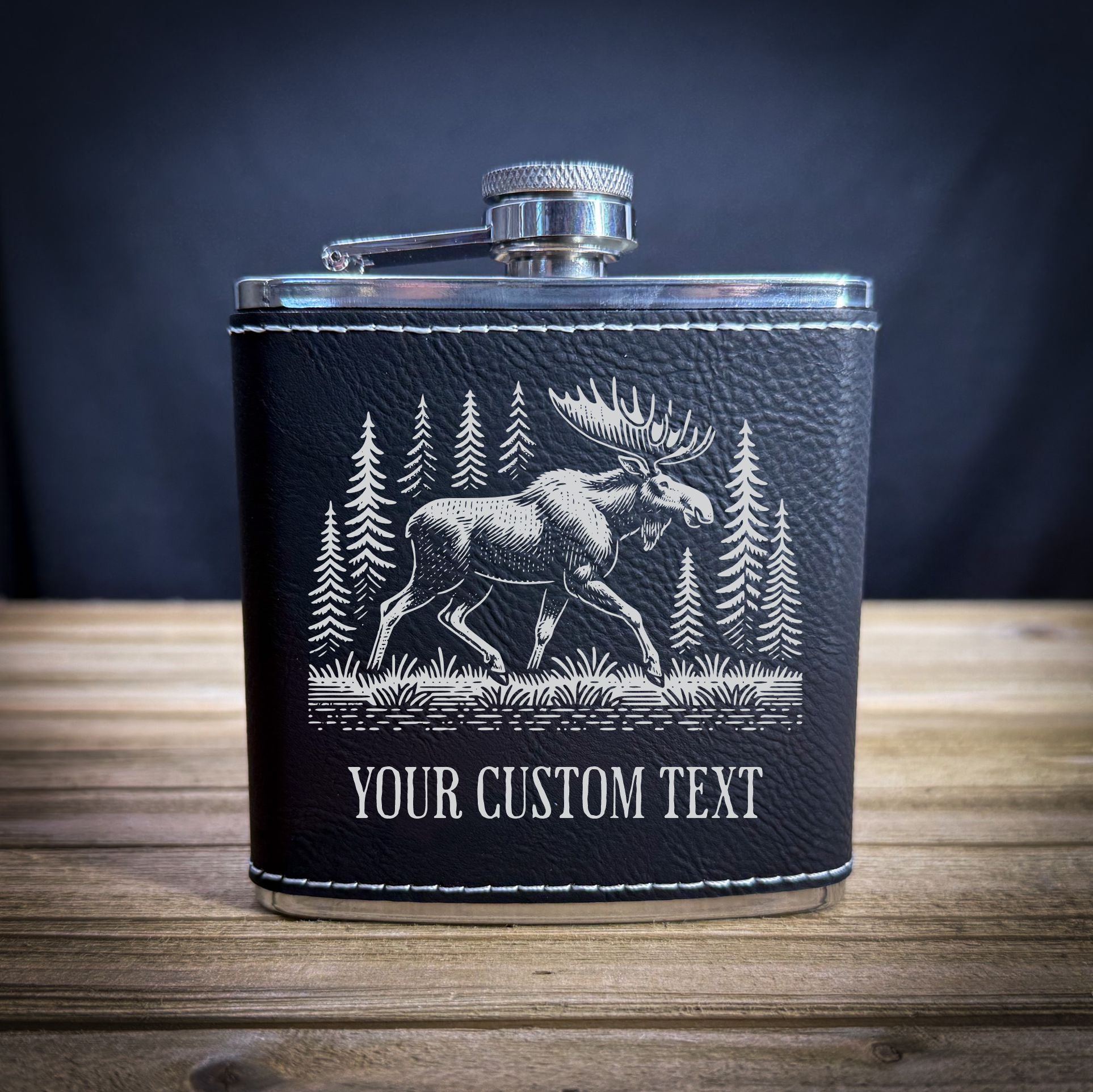 A black leather flask with a silver cap and a white illustration of a moose in a forest setting.