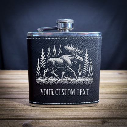 A black leather flask with a silver cap and a white illustration of a moose in a forest setting.