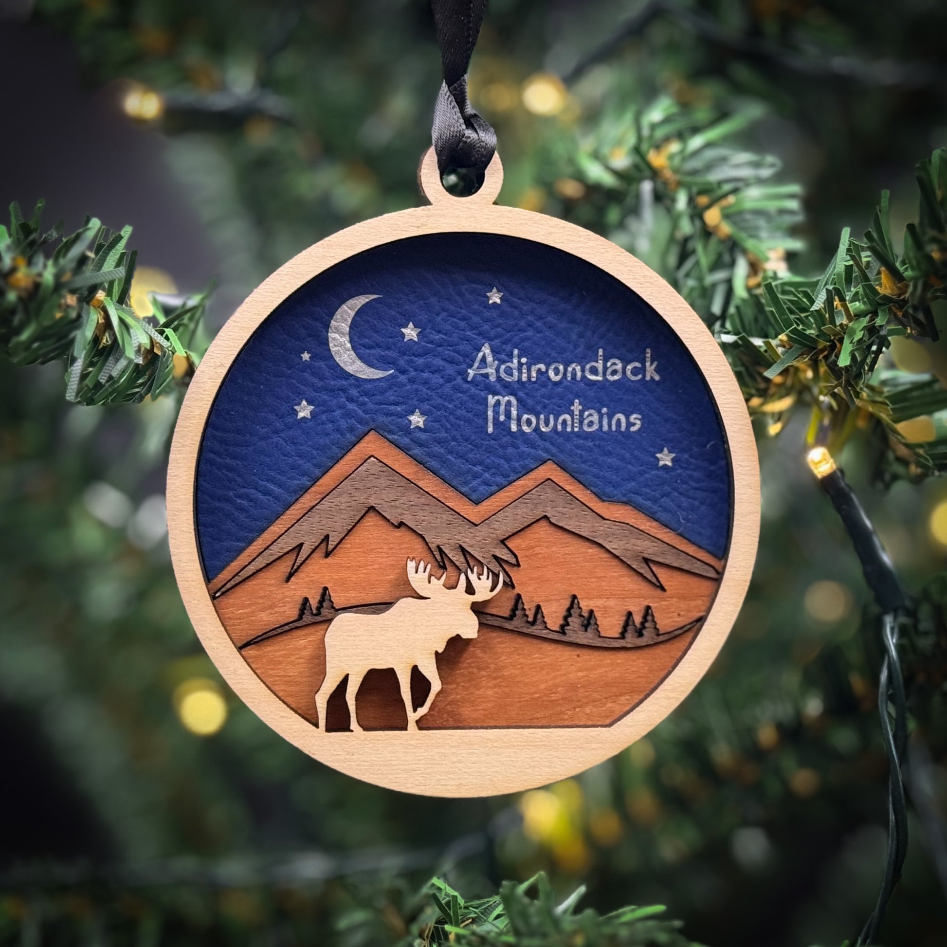 A wooden ornament hanging on a Christmas tree, depicting a moose silhouette against a night sky with mountains and stars.