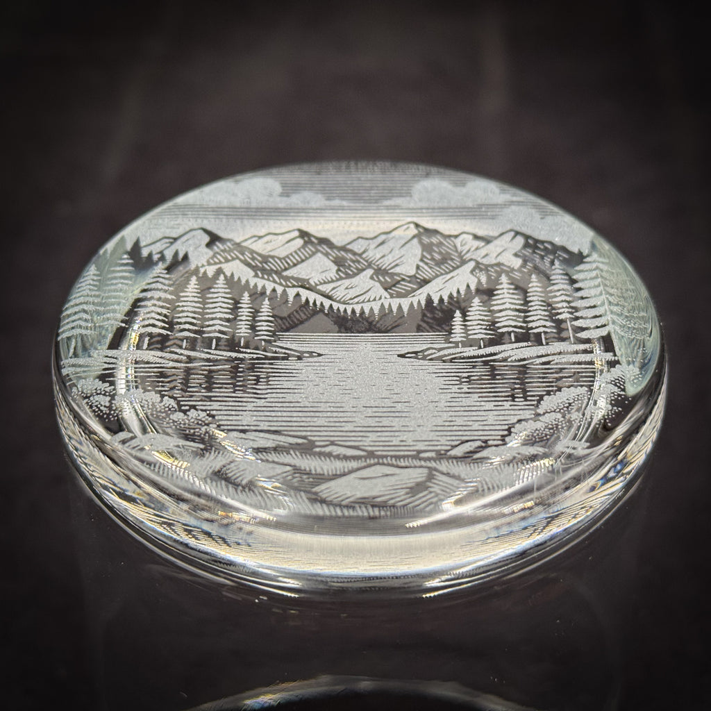 a glass object with a detailed engraved landscape scene depicting mountains, trees, and a lake.