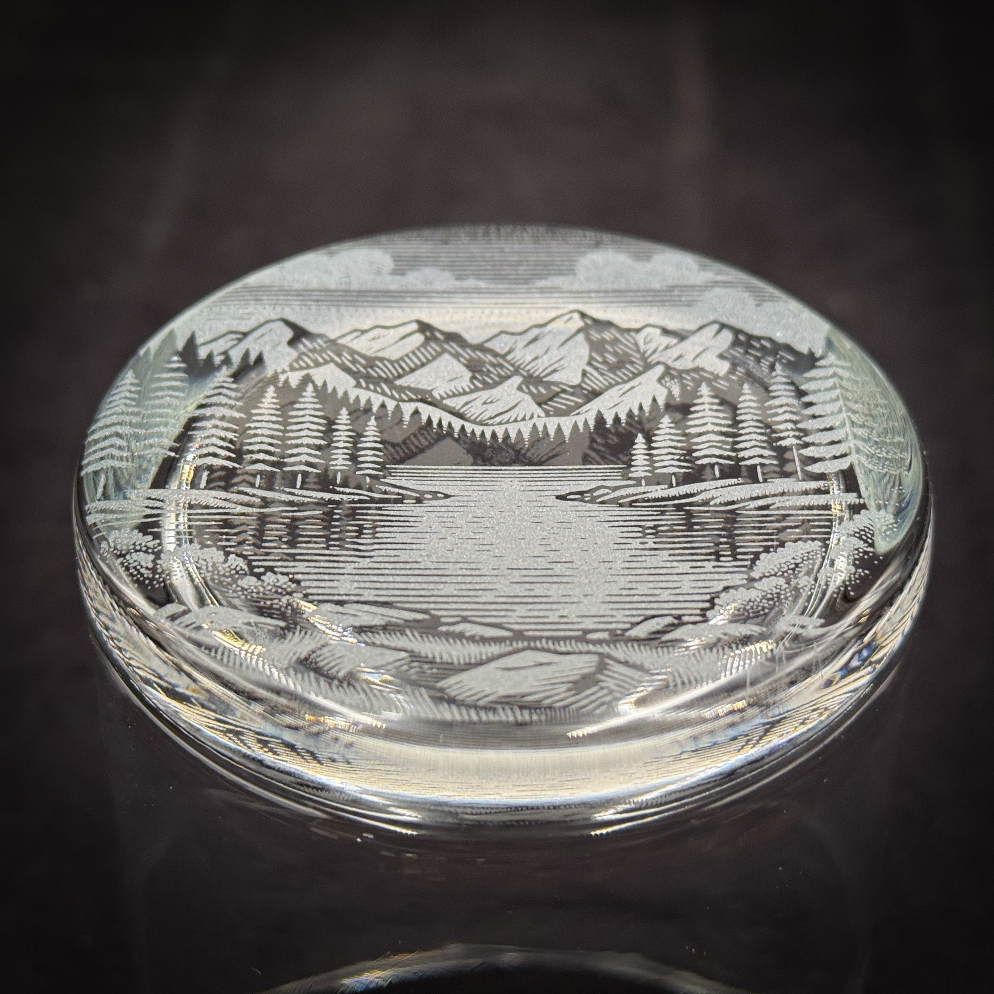 a glass object with a detailed engraved landscape scene depicting mountains, trees, and a lake.