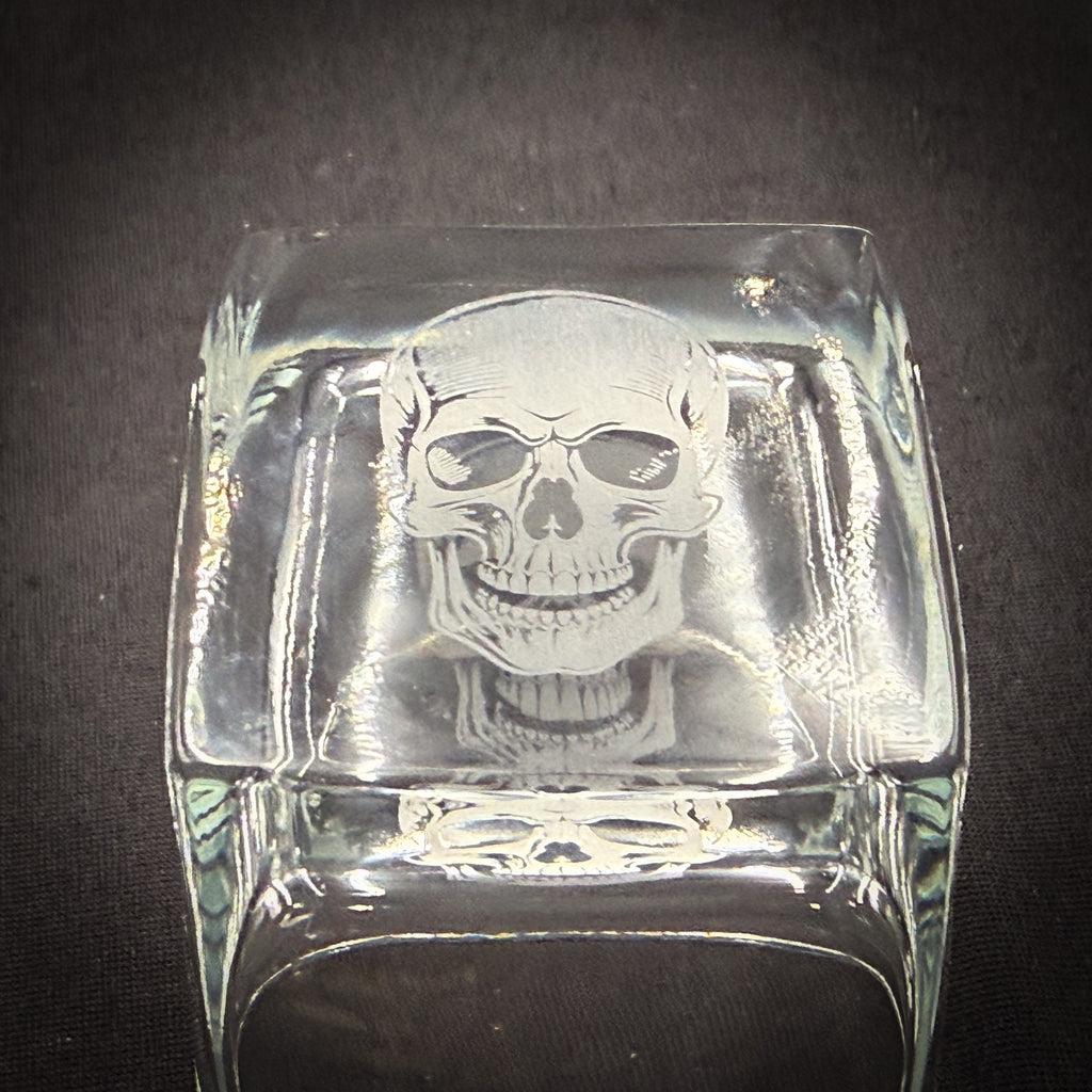 A clear glass object with a skull etched onto it, sitting on a dark surface.