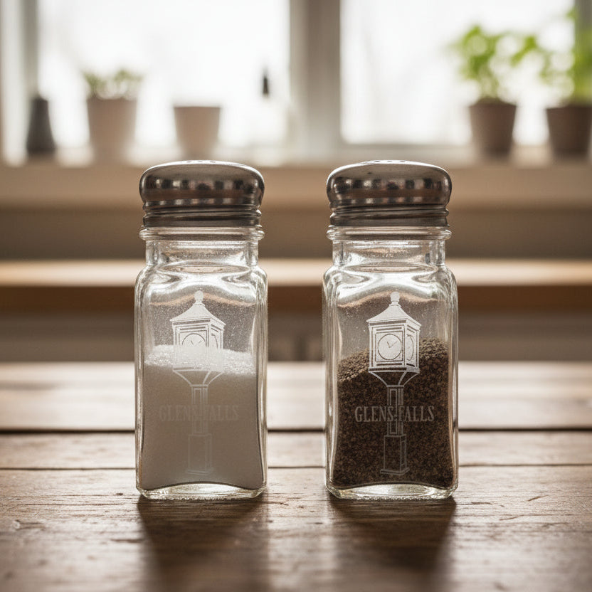 Two glass salt shakers with metal lids on a dark surface and background.