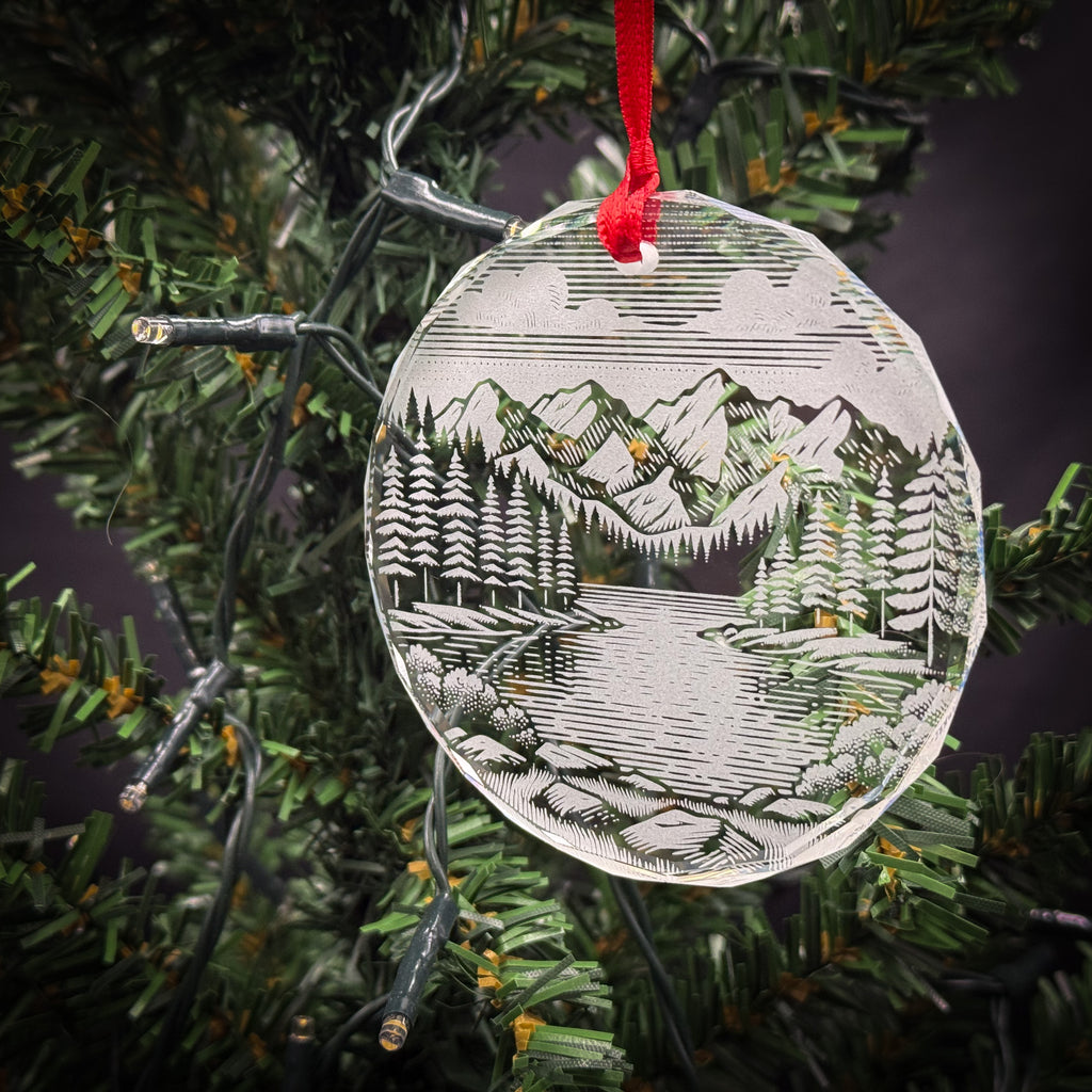 A clear glass ornament with a landscape scene hangs from a Christmas tree, featuring a snowy mountain landscape with pine trees and a lake.
