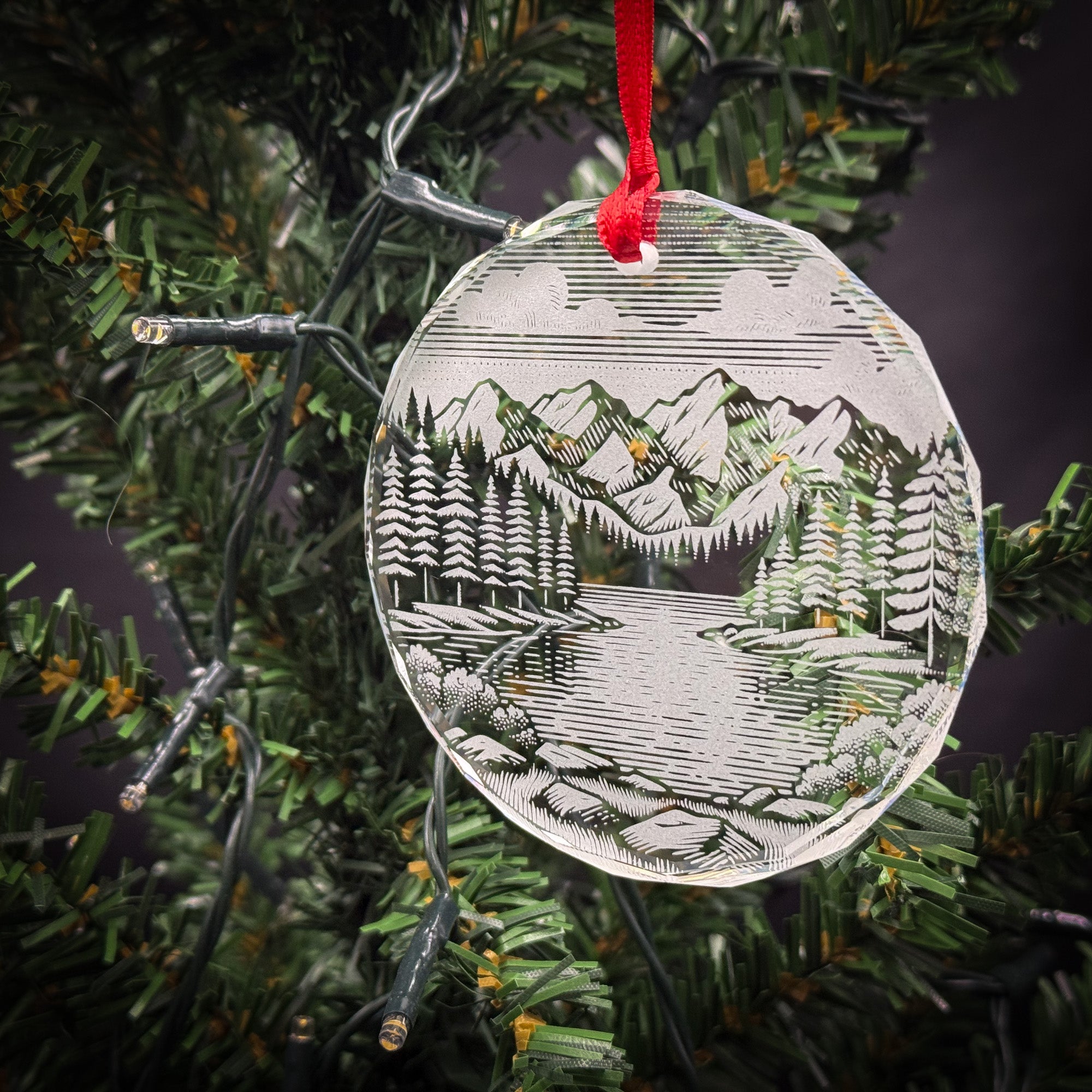 A clear glass ornament with a landscape scene hangs from a Christmas tree, featuring a snowy mountain landscape with pine trees and a lake.