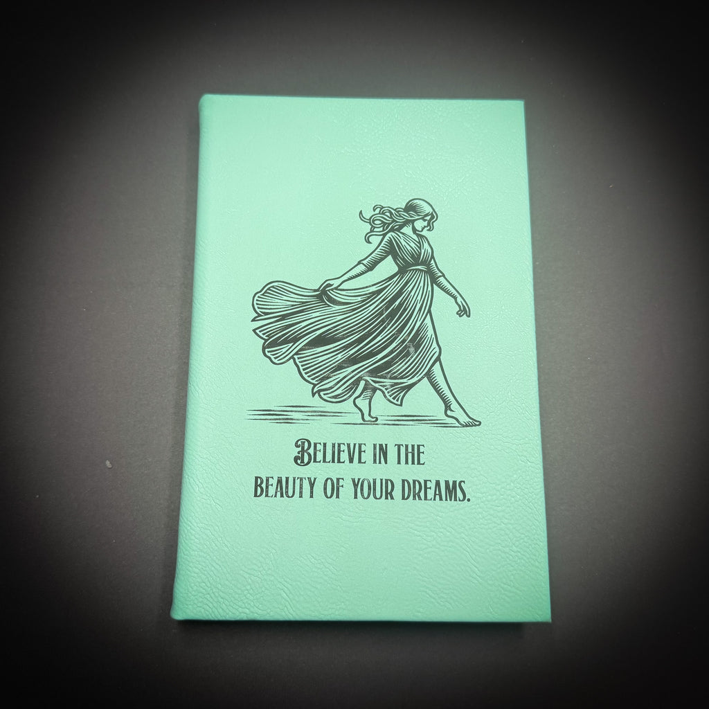 A mint green notebook with a black and white illustration of a woman in a flowing dress and the text "Believe in the beauty of your dreams" printed on it.