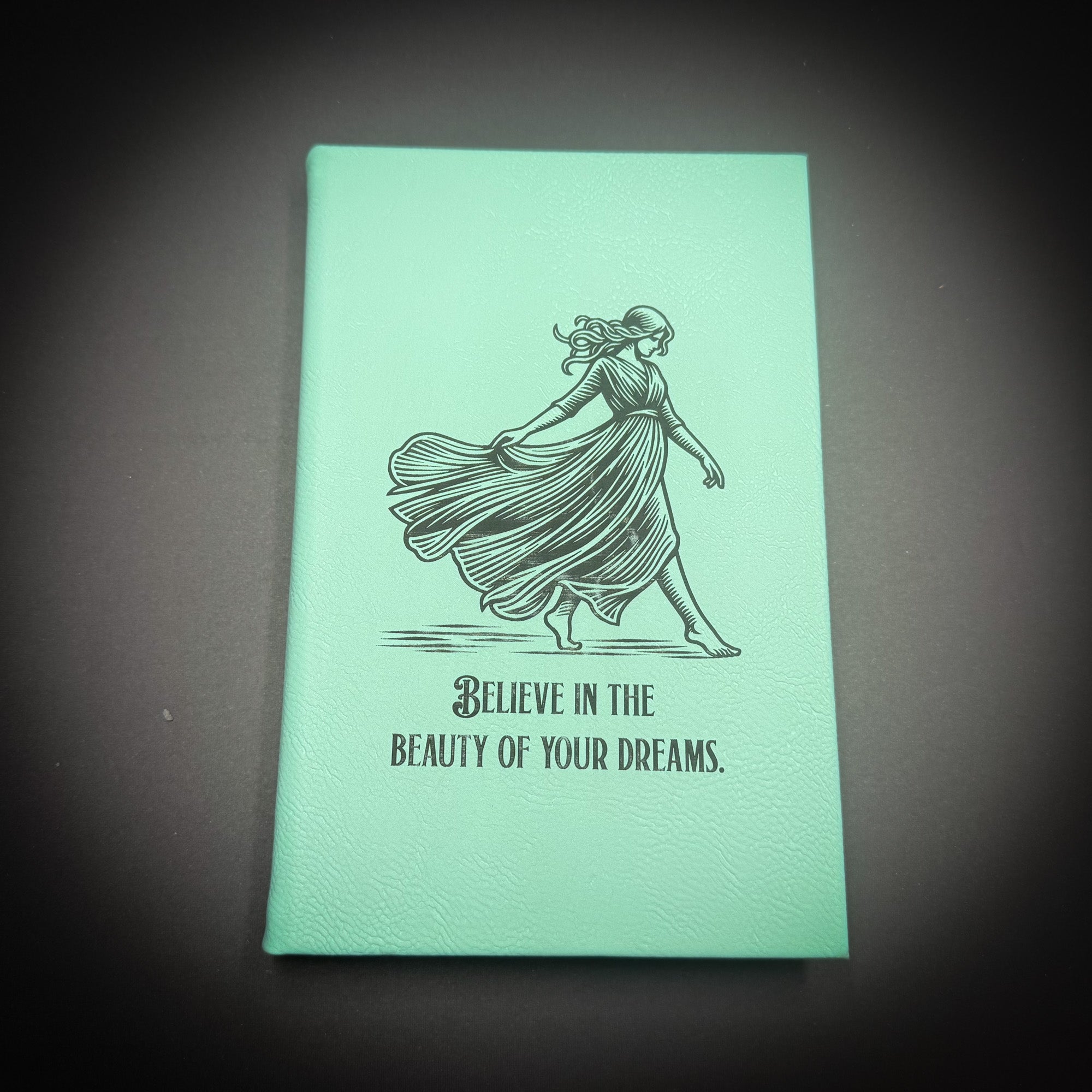 A mint green notebook with a black and white illustration of a woman in a flowing dress and the text "Believe in the beauty of your dreams" printed on it.
