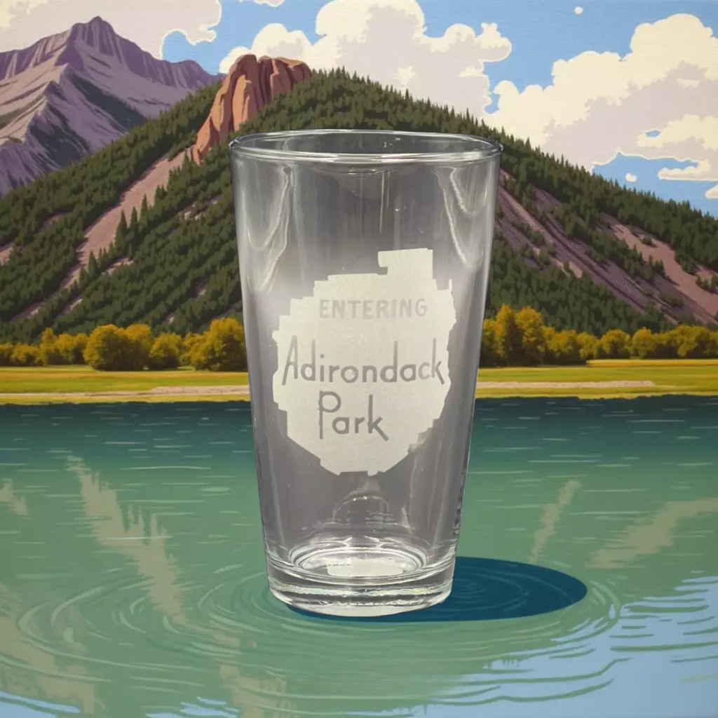 A clear glass with the text "ENTERING Adirondack Park" printed on it, placed on a body of water with mountains in the background.