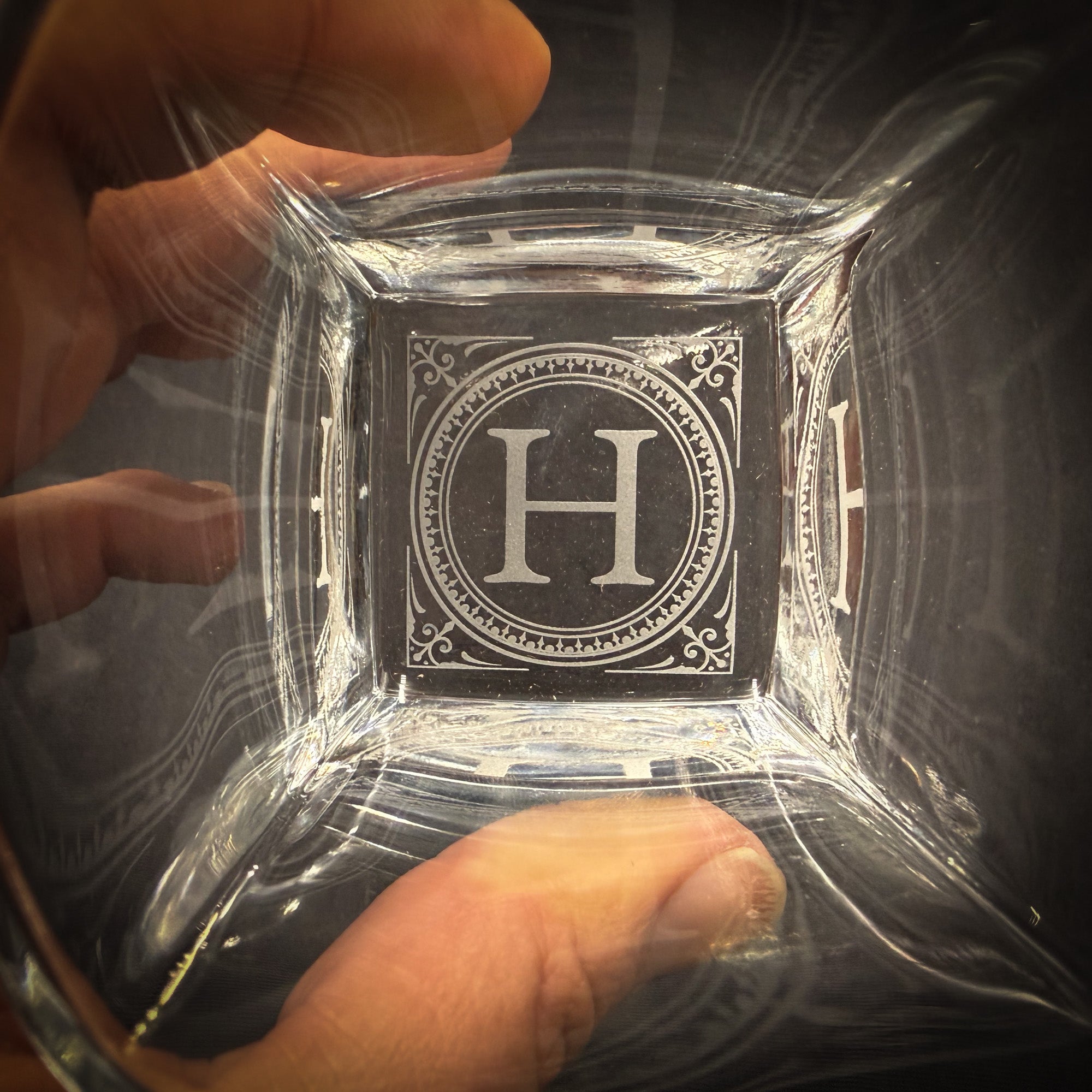 A hand holding a clear glass with a large letter "H" engraved on it.