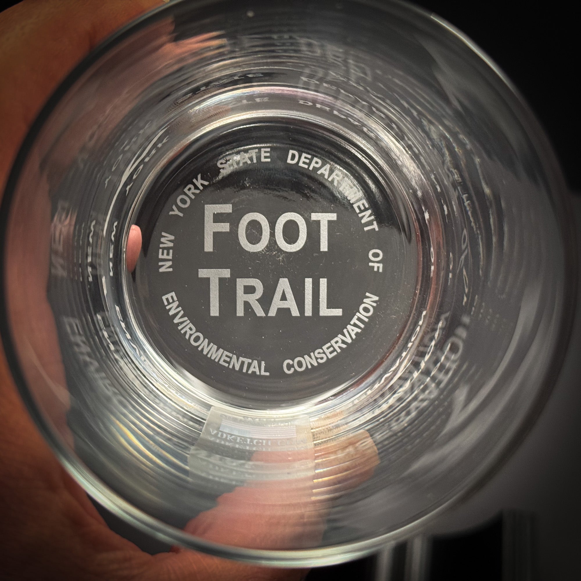 A hand holding a clear glass with the text "FOOT TRAIL" printed on it.