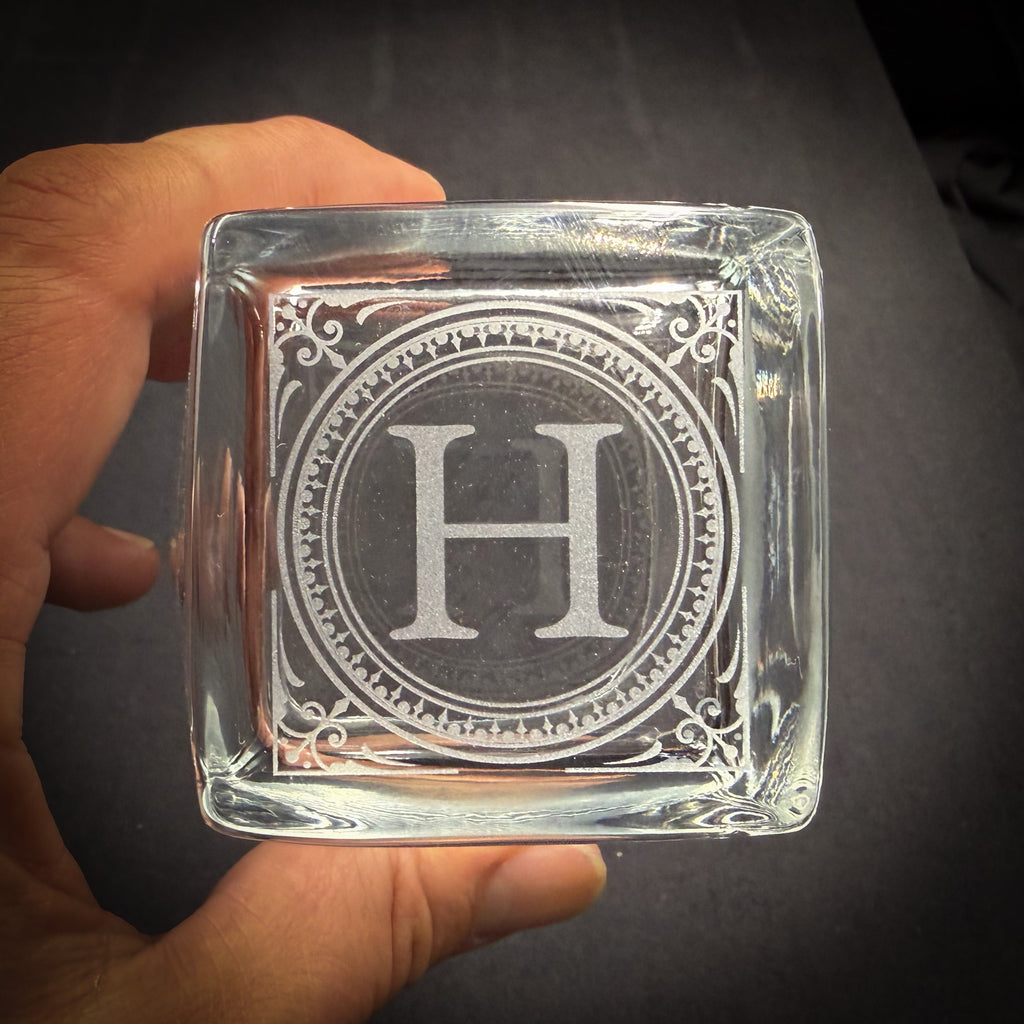 A person's hand holding a clear glass object with an engraved letter "H" in the center.