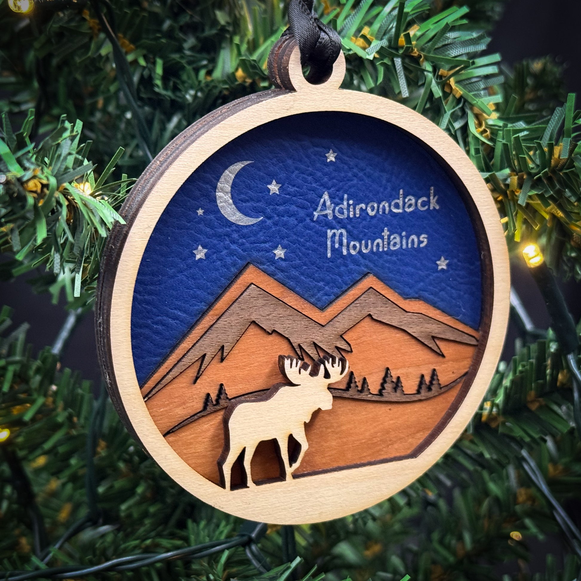 A wooden ornament hanging on a Christmas tree, depicting a moose silhouette against a night sky with mountains and a crescent moon.