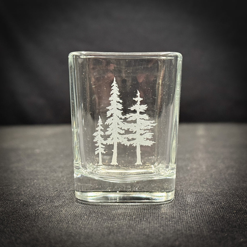 A clear glass shot glass with a white tree design etched onto it, sitting on a dark surface.