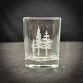 A clear glass shot glass with a white tree design etched onto it, sitting on a dark surface.