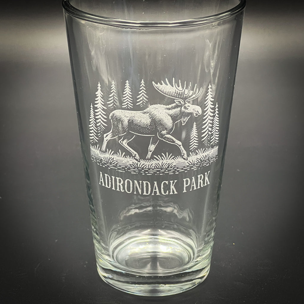 A clear glass tumbler with an etched image of a moose and pine trees on it, along with the text "ADIRONDACK PARK".