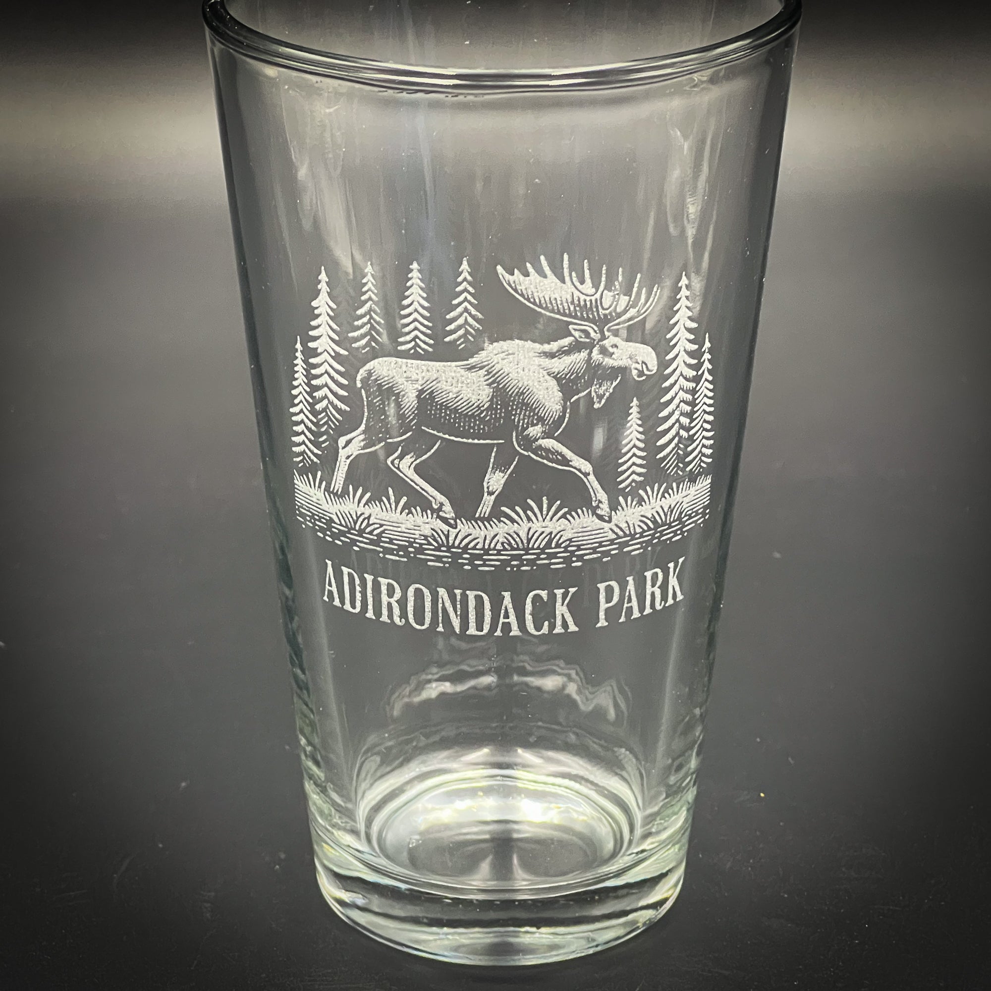 A clear glass tumbler with an etched image of a moose and pine trees on it, along with the text "ADIRONDACK PARK".