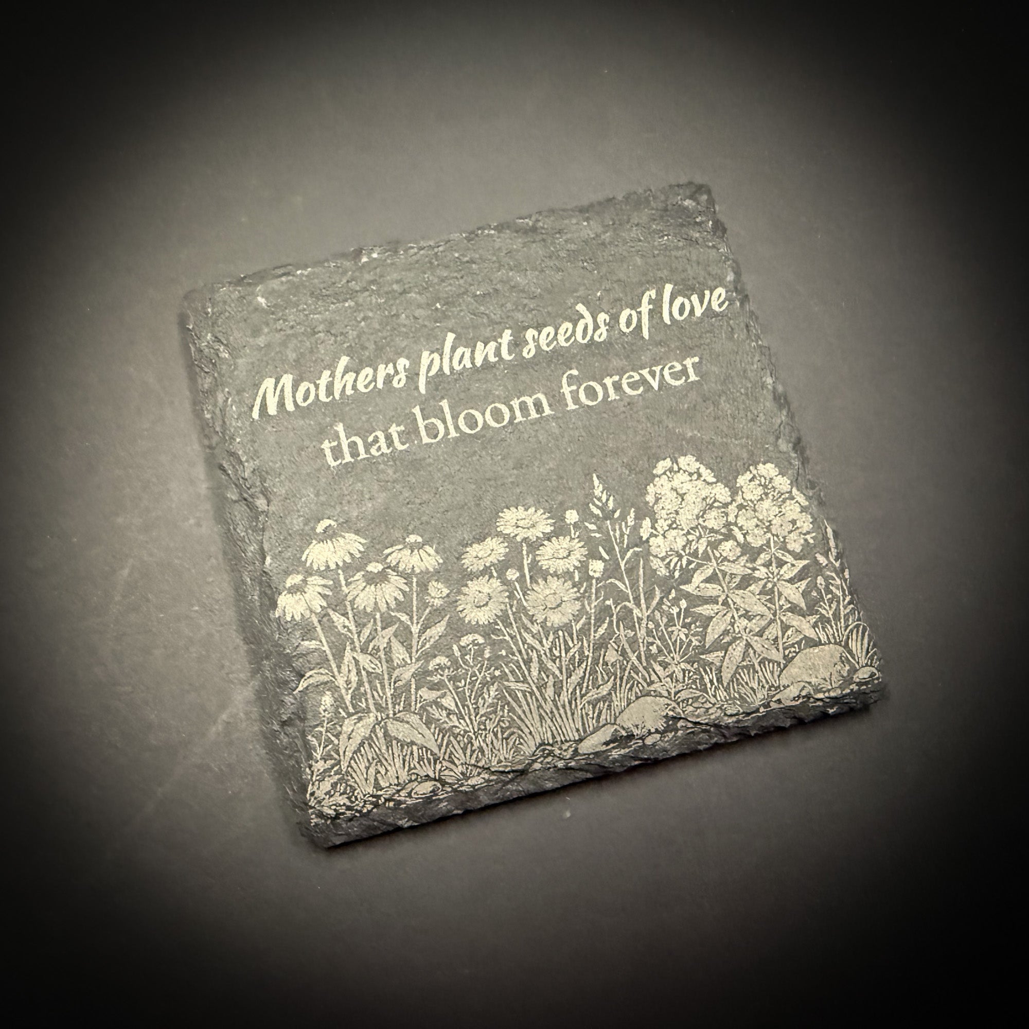 A square stone with a floral design and the text "Mothers plant seeds of love that bloom forever" engraved on it.
