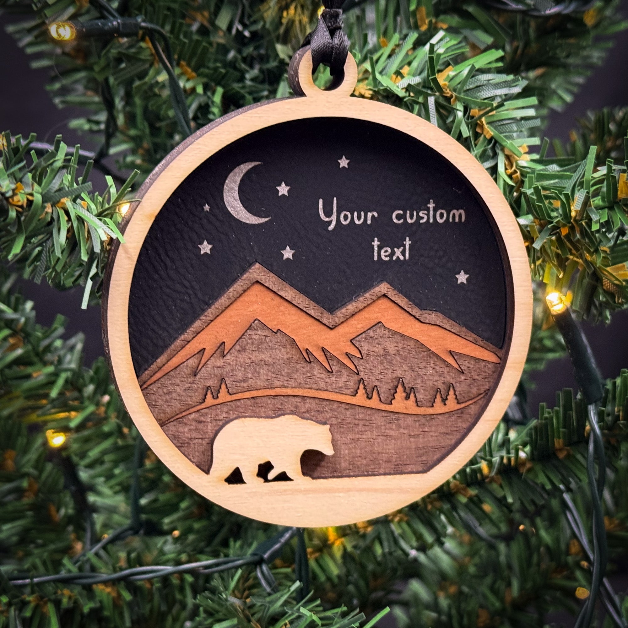 A wooden ornament hanging on a Christmas tree, depicting a bear silhouette against a night sky with mountains and stars.