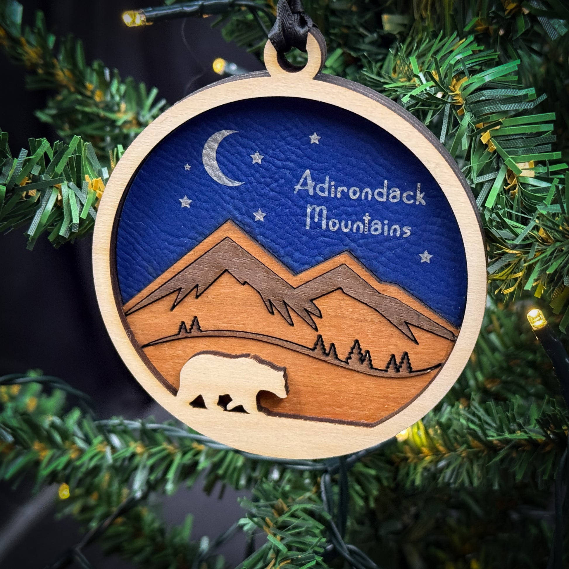 A wooden ornament hanging on a Christmas tree, depicting a bear silhouette against a night sky with mountains and stars.