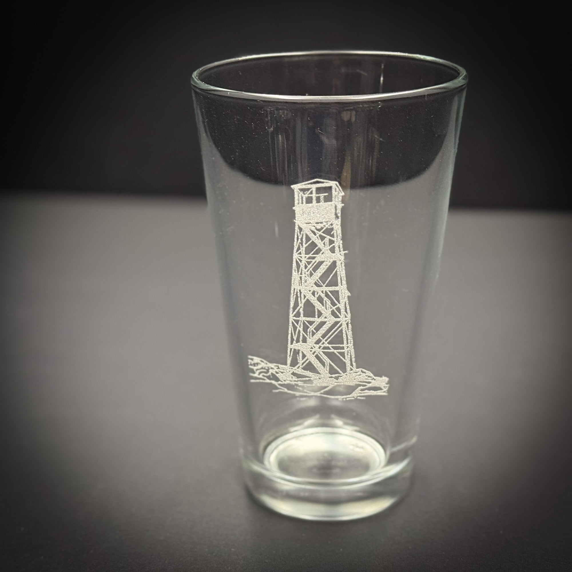 A clear glass with a white lighthouse design etched on it, sitting on a dark surface.