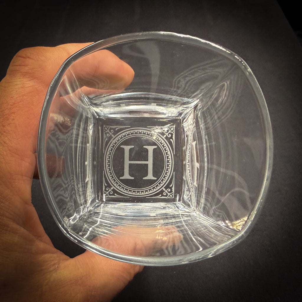 A hand holding a clear glass with a large letter "H" engraved on it.